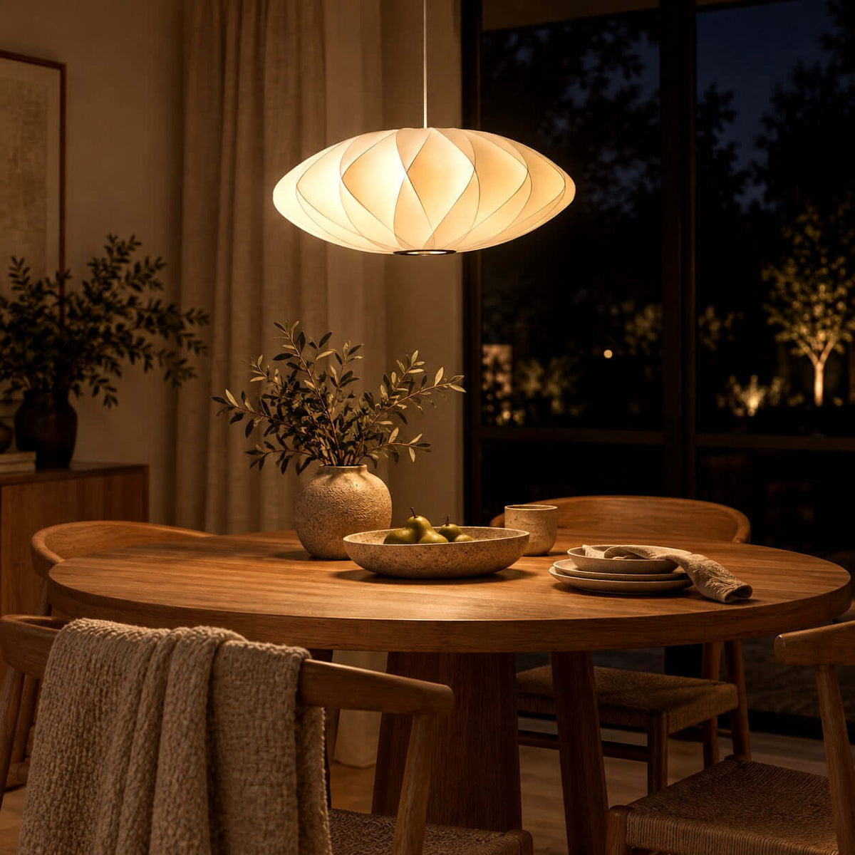 Corvya silk pendant light glowing warmly above a round wood dining table beside dark windows at night.