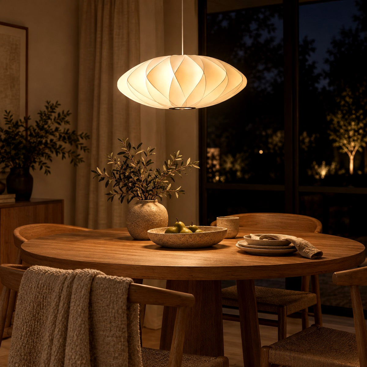 Corvya silk pendant light glowing warmly above a round wood dining table beside dark windows at night.